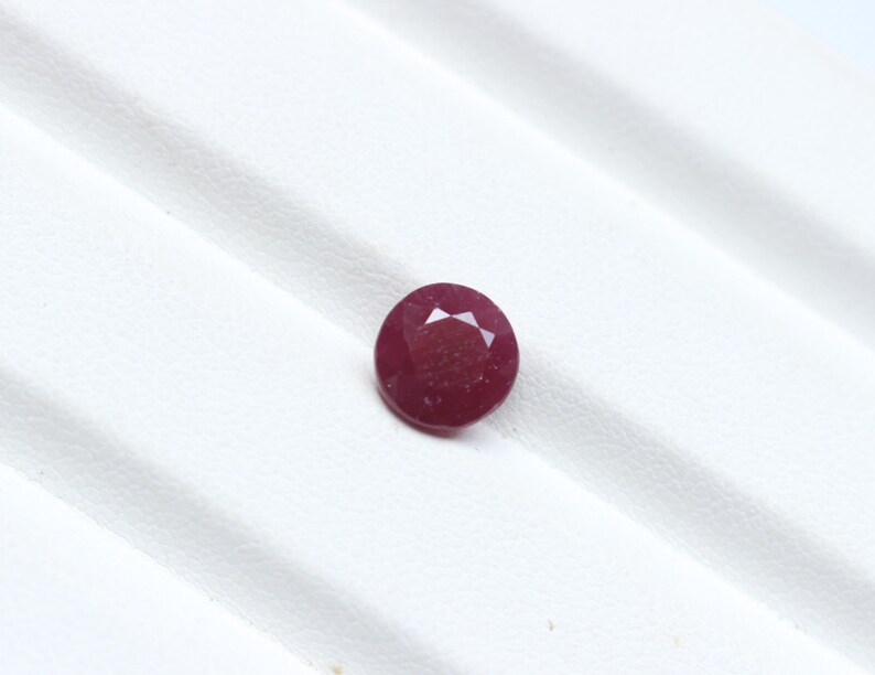 K&ouml;nnte beinhalten: Ein runder, facettierter Rubin-Edelstein mit tiefroter Farbe. Der Edelstein ist vor einem wei&szlig;en Hintergrund platziert, was seine Farbe und Klarheit hervorhebt. Die Facetten des Rubins fangen das Licht ein und erzeugen einen funkelnden Effekt. Geeignet f&uuml;r die Schmuckherstellung.