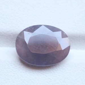 Op de afbeelding: Een gefacetteerde, ovale edelsteen met een gedempte paarse tint. Het gepolijste oppervlak heeft meerdere facetten die licht reflecteren, waardoor een sprankelend effect ontstaat. De steen is geplaatst tegen een witte achtergrond.