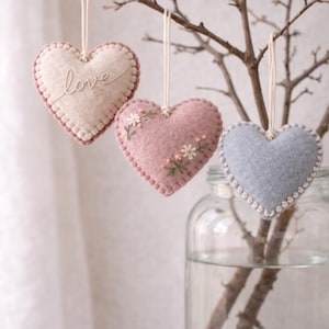 May include: Three heart-shaped ornaments hanging from a branch. The ornaments are made of felt in cream, pink, and blue. The cream-colored heart has the word "love" embroidered on it. The pink heart has embroidered flowers. The blue heart has a stitched border.