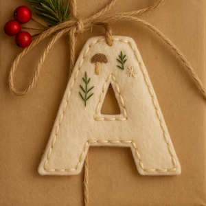May include: A cream-colored felt letter "A" with embroidered forest-themed details, including a mushroom, leaves, and a star. The letter is attached to a wrapped package with twine and decorated with a sprig of evergreen and red berries.