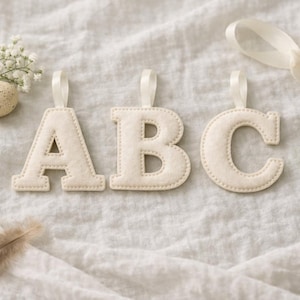 May include: Three off-white fabric letters A, B, and C, each featuring a small fabric loop at the top for hanging. The letters are arranged horizontally and are accompanied by a small speckled egg, white flowers, a feather, and a white ribbon.