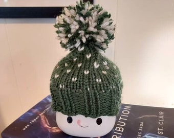 Könnte beinhalten: Ein weißer Keramiktopf mit einem lächelnden Gesicht, bedeckt mit einer grün-weißen Strickmütze mit Bommel. Der Topf steht auf einem Buch mit dem Titel "Retribution" von Scarlett St. Clair.