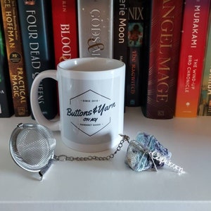 May include: A white ceramic mug with a black logo that says "Buttons & Yarn Oh My! Handknit Goods" and a silver tea infuser with a chain attached to a blue and green crystal with a silver tag that says "Capricorn".