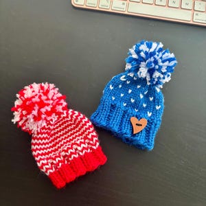 May include: Two miniature knitted hats with pom-poms. One is red and white, the other is blue with white accents and a wooden heart. The hats are handmade and likely for a doll or small pet. They are placed on a dark surface near a keyboard.