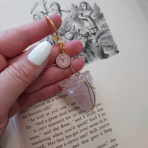 May include: A gold key chain with a pink clock charm and a pink crystal attached to it. The crystal is wrapped with gold wire. The key chain is held in a hand over a book with the text "The Garden of Live Flowers" and a black and white illustration of Alice in Wonderland.
