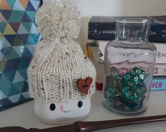 May include: A white ceramic mug with a knitted hat and a brown heart on the side. The mug is sitting on a table with a wooden wand and a glass jar filled with green dice.