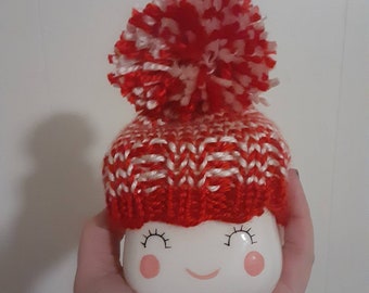 Könnte beinhalten: Eine weiße Keramiktasse mit einem lächelnden Gesicht, Rouge und Wimpern, bedeckt mit einer rot-weiß gestrickten Mütze mit Bommel. Die Tasse wird in einer Hand gehalten.