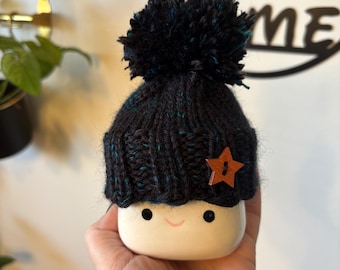 Könnte beinhalten: Eine weiße Keramiktasse mit einem lächelnden Gesicht, die eine winzige schwarze Strickmütze mit Bommel und einem Holzstern trägt. Die Tasse wird in einer Hand gehalten, mit einem verschwommenen Hintergrund mit den Worten "Sweet Home".