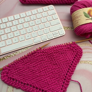 May include: A pink knitted dishcloth with a wooden knitting needle and a ball of pink yarn. The dishcloth is in the shape of a triangle and has a scalloped edge.