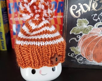 May include: A white ceramic mug with a smiling face, wearing an orange and white striped knitted hat with a white pom-pom and a brown star. The mug is sitting on a shelf with books behind it.