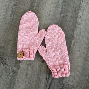 Puede incluir: Un par de mitones de punto rosa con lunares blancos. Los mitones tienen un botón en uno de ellos.