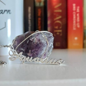 May include: A silver necklace with a purple amethyst crystal wrapped in wire. The pendant is labeled "Aquarius".