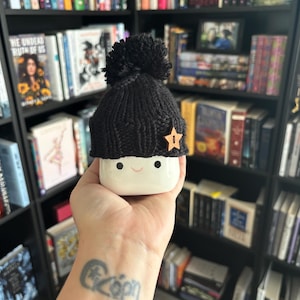 May include: A white ceramic mug with a happy face and a black knitted hat with a pom-pom and a gold star on top.