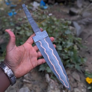 A person holds an unfinished Damascus steel knife blade with a wavy copper stripe across its center.