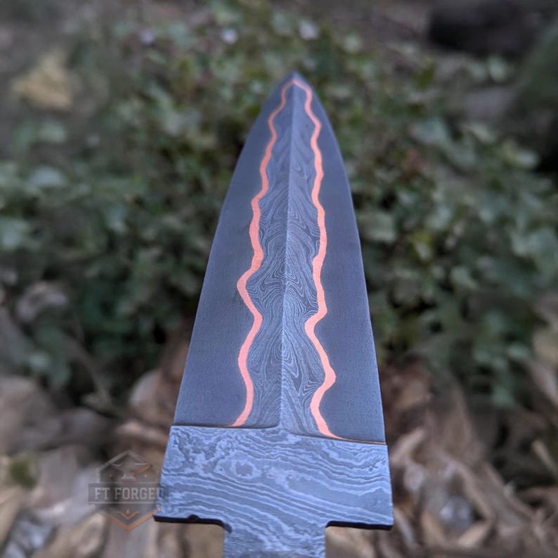A close-up view of the tip and pattern of a Damascus steel knife blade with wavy copper inlays.