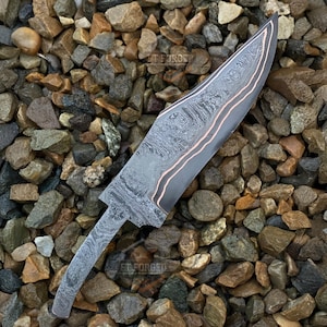 May include: A Damascus steel knife blade with a long, pointed shape. The blade features a dark, patterned finish with a copper-colored accent line along the edge. The knife is set against a background of small, multicolored stones.