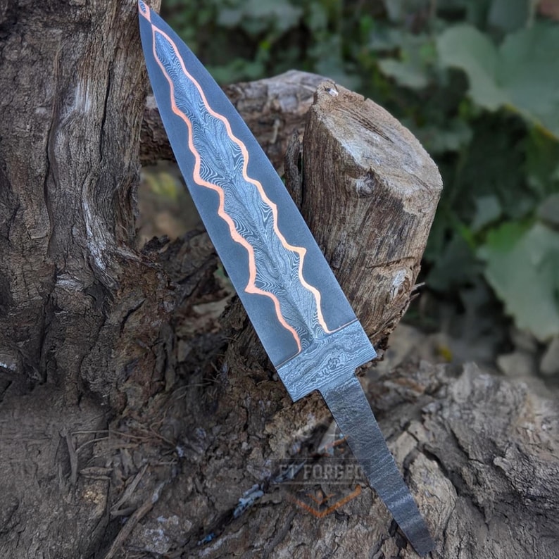 A Damascus steel knife blank with a wavy copper core rests upright against a tree stump.