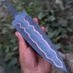 A person holds a Damascus steel knife blank with a wavy copper accent, showing the tip pointing downward.