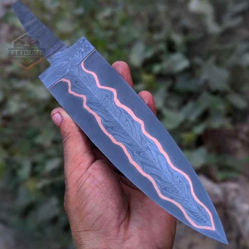 A person holds a Damascus steel knife blank with a wavy copper accent, showing the tip pointing downward.