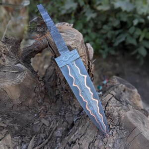 A Damascus steel knife blank with a copper stripe rests vertically on a cut log in an outdoor setting.