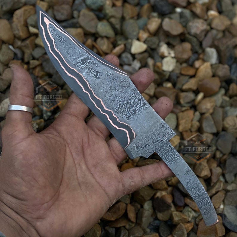 May include: A Damascus steel knife blade with a copper-colored accent. The blade has a textured pattern and is held in a hand. The text "FT FORGED" is visible on the blade. The knife is designed for crafting or collecting.