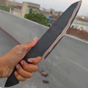 Könnte beinhalten: Ein schwarzes Messer mit einem geschichteten Design und silbernen und orangefarbenen Akzenten. Das Messer wird in einer Hand gehalten, mit dem Text "FT FORGED" sichtbar. Die Klinge hat eine lange, spitze Form.