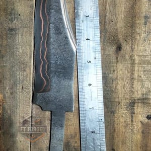 May include: A forged steel knife blade with a copper-colored accent, approximately 10 inches long, next to a ruler. The blade has a textured pattern and a curved edge. The knife is on a wooden surface, with the text "FT FORGED" visible.