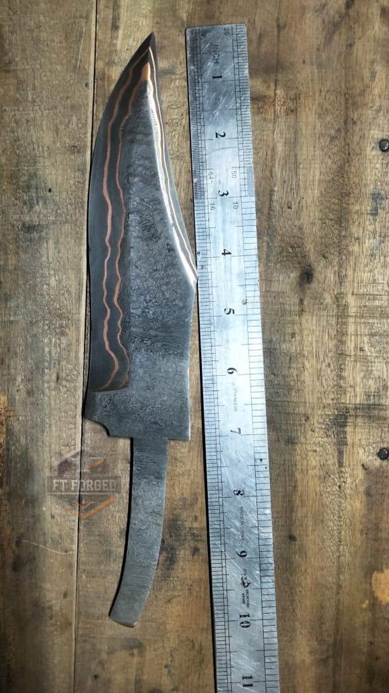 May include: A forged steel knife blade with a copper-colored accent, approximately 10 inches long, next to a ruler. The blade has a textured pattern and a curved edge. The knife is on a wooden surface, with the text "FT FORGED" visible.