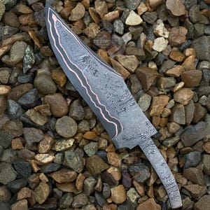 May include: A Damascus steel knife with a curved blade and a copper-colored edge. The blade has a unique pattern and the text "FT FORGED" is visible. The knife is displayed on a bed of small rocks.