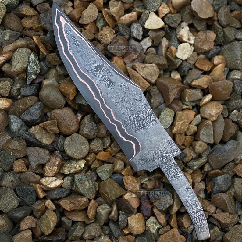 May include: A Damascus steel knife with a curved blade and a copper-colored edge. The blade has a unique pattern and the text "FT FORGED" is visible. The knife is displayed on a bed of small rocks.