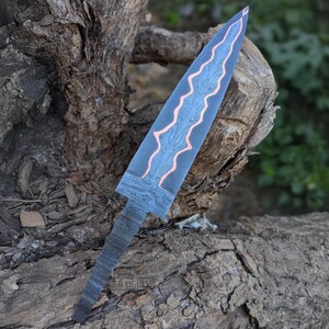 A Damascus steel knife blank featuring a wavy copper inlay lies flat on a tree stump.