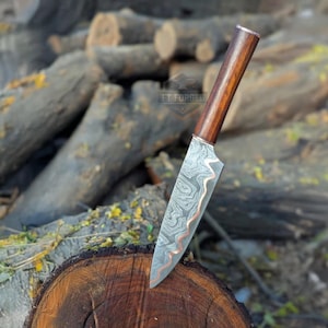 May include: A Damascus steel knife with a wooden handle and copper detailing. The blade has a distinctive swirling pattern. The knife is shown on a wooden surface, with a blurred background of logs.