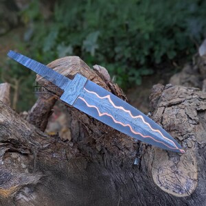 A horizontal view of a Damascus steel knife blank resting on a textured wooden stump outdoors.