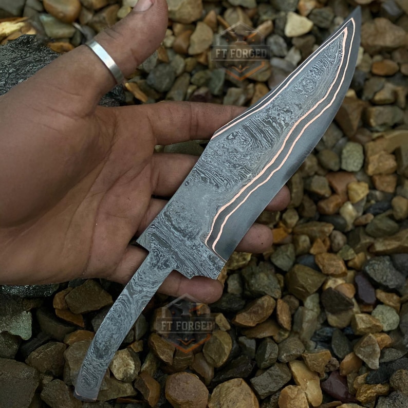 May include: A Damascus steel knife blade with a patterned design and copper-colored accents. The blade is held in a hand, and the text "FT FORGED" is visible on the blade.