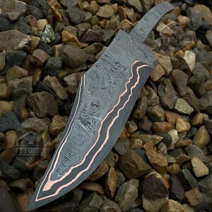 May include: A forged steel knife blade with a patterned design and copper-colored accents. The blade has a curved edge and a textured surface. The knife is displayed on a bed of small, multicolored stones. The text "FT FORGED" is visible.