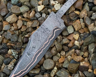 May include: A large, Damascus steel knife with a patterned blade and a copper-colored accent along the edge. The knife is lying on a bed of small, multicolored stones, showcasing its intricate design and craftsmanship.