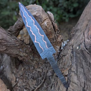 A Damascus steel knife blank with a copper inlay is positioned vertically against tree wood.