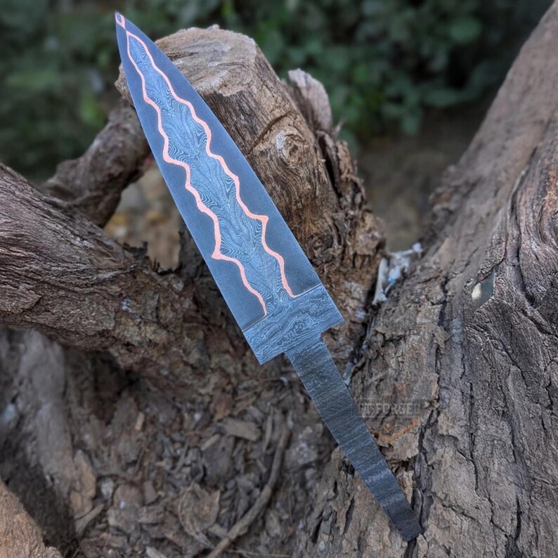 A Damascus steel knife blank with a copper inlay is positioned vertically against tree wood.