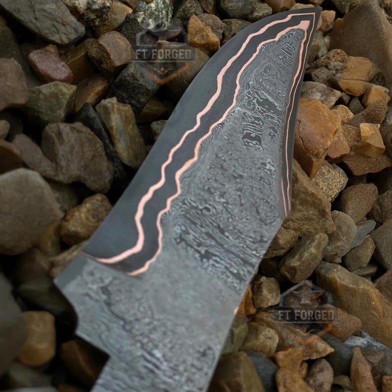 May include: Close-up of a forged knife blade with a unique pattern. The blade has a dark gray finish with a wavy copper-colored edge. The blade is set against a background of small, multicolored rocks. The text "FT FORGED" is visible.