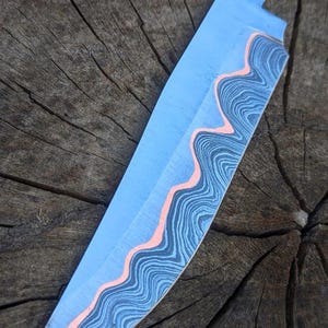 Op de afbeelding: Een op maat gemaakt, blauw koksmes met een uniek golfpatroon langs de rand van het lemmet. Het lemmet heeft een gelaagd ontwerp met tinten blauw, zwart en een zalmkleurig accent. Het mes ligt op een rustieke houten achtergrond.
