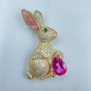 May include: A gold-coloured bunny brooch encrusted with clear crystals, featuring pink-painted inner ears and a green eye. The bunny holds a large, pear-shaped, magenta-coloured gemstone. The brooch is a piece of jewellery.