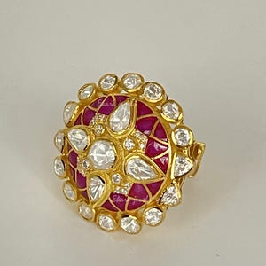 Puede incluir: Un anillo de oro con un diseño grande y redondo. El anillo presenta un patrón central de piedras preciosas rojas rodeadas de piedras preciosas blancas claras en forma de lágrima y redondas. El anillo tiene una banda de oro y está sobre un fondo claro.