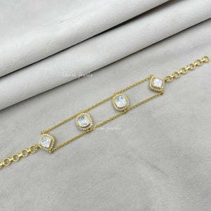 Könnte beinhalten: Goldfarbenes Armband mit vier klaren, quadratischen und ovalen Steinen, die jeweils von kleineren, klaren Steinen umgeben sind. Das Armband hat ein filigranes Kettendesign und wird auf einer hellgrauen Oberfläche präsentiert. Der Markenname "Elara Jewels" ist sichtbar.