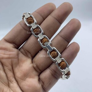 Op de afbeelding: Een zilveren schakelarmband met afwisselende rechthoekige schakels met een gestippeld textuurpatroon en kleine, ronde, bruine, natuurlijk getextureerde rudraksha-kralen in elke schakel, afgewerkt met een gepolijste metalen glans.