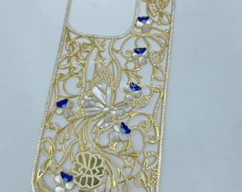 以下が含まれることがあります： 金色の携帯電話ケース。精巧な透かし彫りのデザイン。蝶と花のモチーフで、白と青の装飾が施されています。小さなキラキラ光るクリスタルがちりばめられています。
