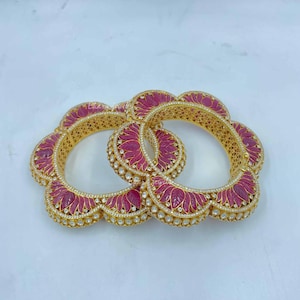Puede incluir: Dos brazaletes dorados con un diseño floral. Los brazaletes presentan una incrustación rosa oscuro en forma de pétalo, acentuada con pequeñas piedras blancas. El intrincado trabajo de filigrana dorada añade detalles a la joyería.