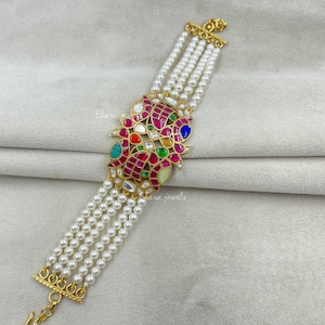 Op de afbeelding: Een armband met meerdere strengen witte parels en een centrale goudkleurige metalen plaat ingelegd met levendige rode, groene, blauwe en gele stenen. Het ontwerp heeft vier rijen parels aan weerszijden en een goudkleurige haaksluiting.