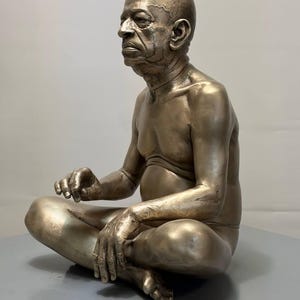 May include: A metallic bronze-toned sculpture of an elderly person with a bald head, seated in a cross-legged position. The figure has a detailed facial expression, visible skin texture, and is depicted with a bare torso and arms resting on legs.