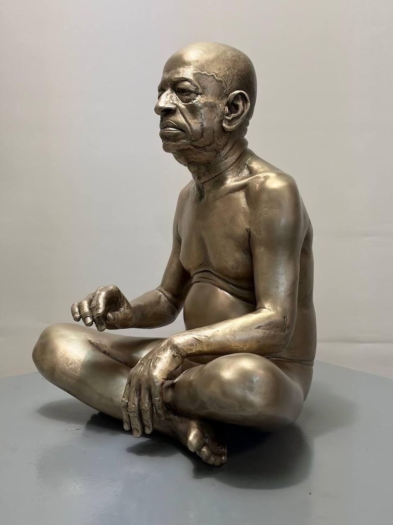 May include: A metallic bronze-toned sculpture of an elderly person with a bald head, seated in a cross-legged position. The figure has a detailed facial expression, visible skin texture, and is depicted with a bare torso and arms resting on legs.