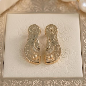 May include: A pair of gold-toned earrings featuring a flat, elongated shape with intricate, symmetrical engraved patterns. Each earring has a circular top section and a wider, curved bottom section, both decorated with fine lines and small dots.